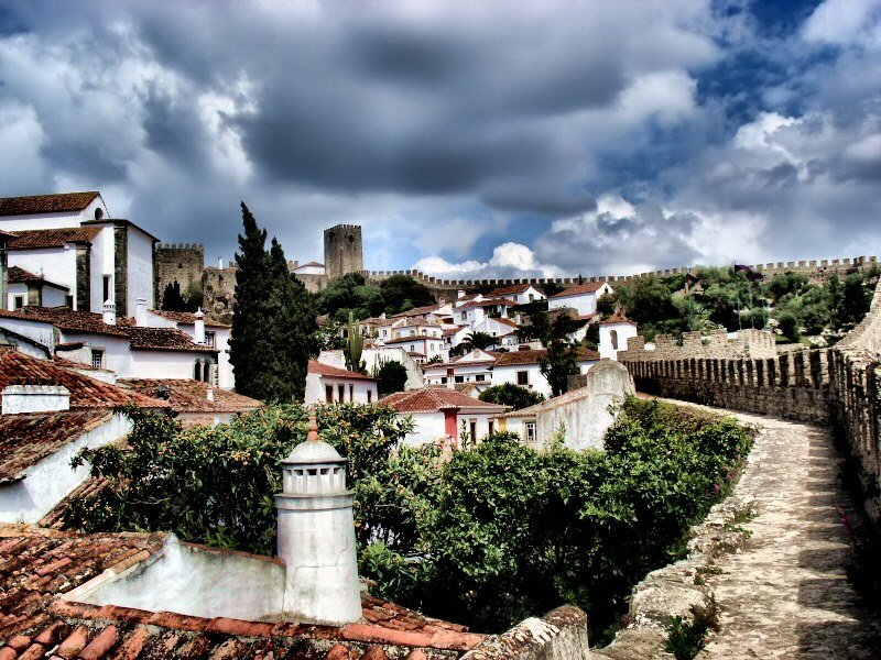 View walking along the stone walls of Obidos overlooking whitewashed rooftops and the castle towers under a dramatic sky