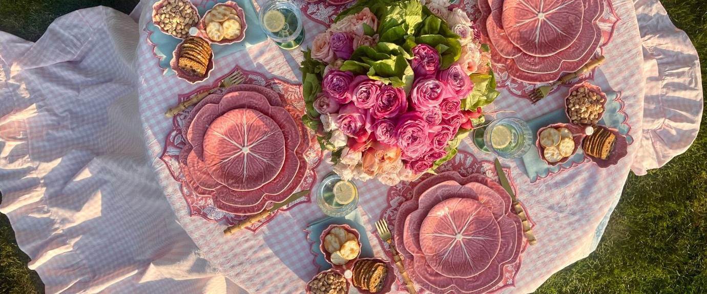 Overhead view of a picnic table set with pink cabbage-leaf Bordallo Pinheiro plates, roses and gingham tablecloth