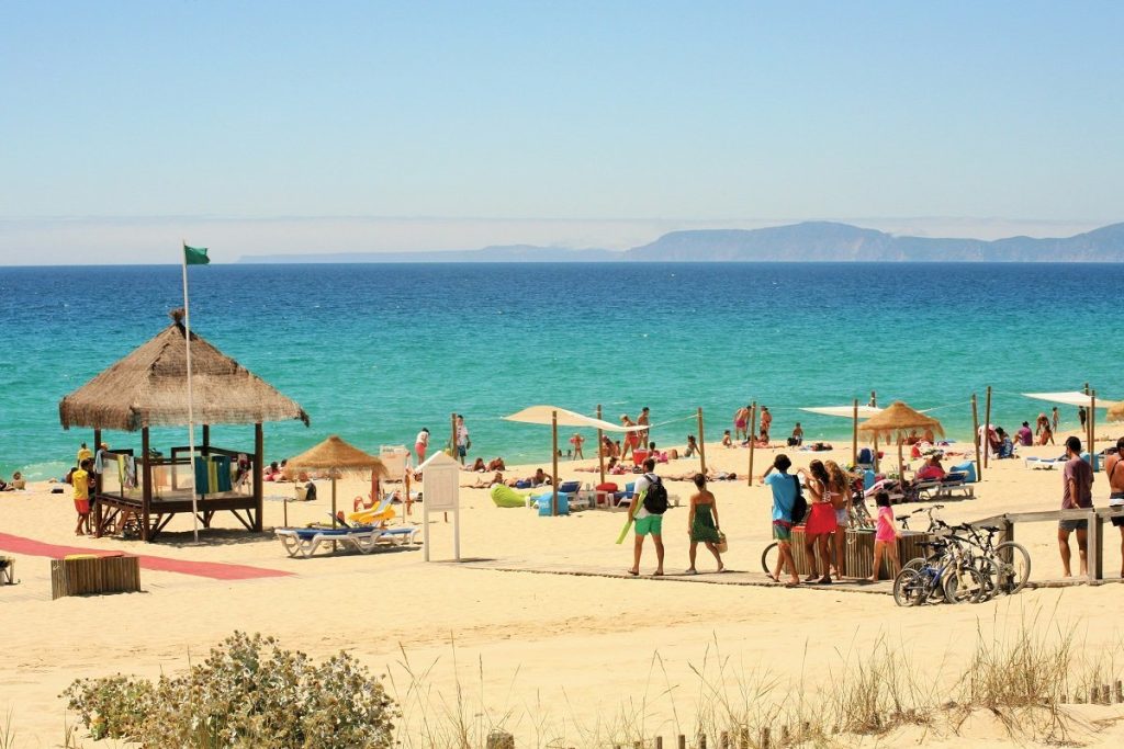 Sunny view of Praia da Comporta beach with turquoise water, a thatched sunshade and people enjoying the sand, Portugal