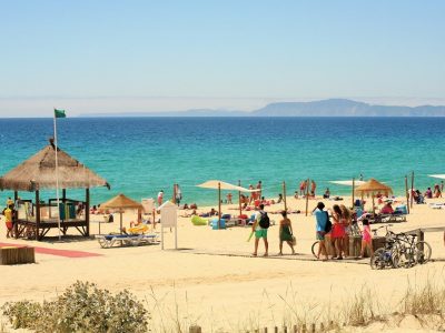 Praia da Comporta, Portugal Sunny view of Praia da Comporta beach with turquoise water, a thatched sunshade and people enjoying the sand, Portugal