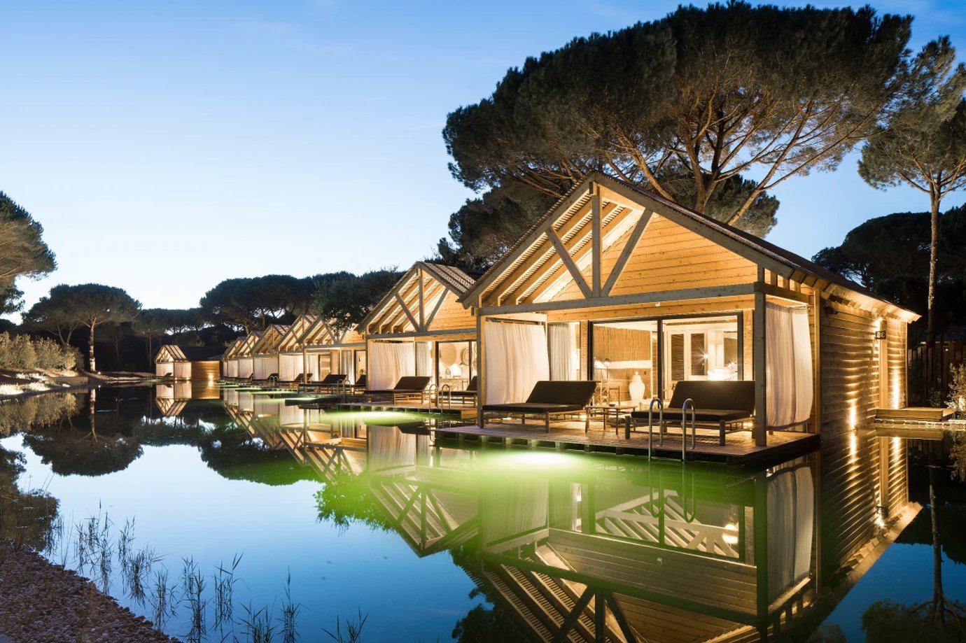 Timber cabana suites reflected in the bio-pool at dusk, Sublime Comporta hotel, Portugal
