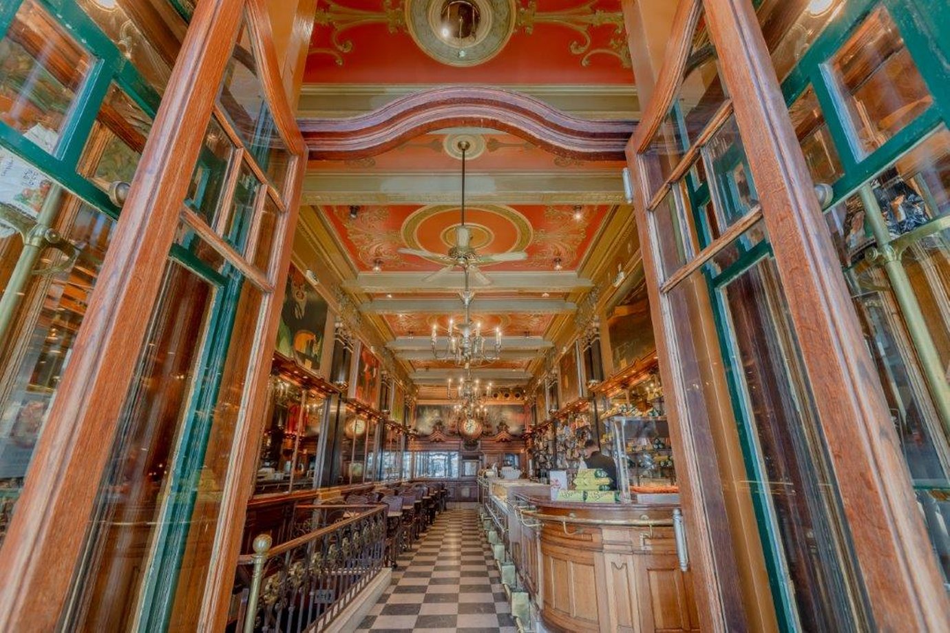 The long interior of A Brasileira in Lisbon seen through open wooden doors, with a painted orange ceiling, chandeliers and a chequered floor