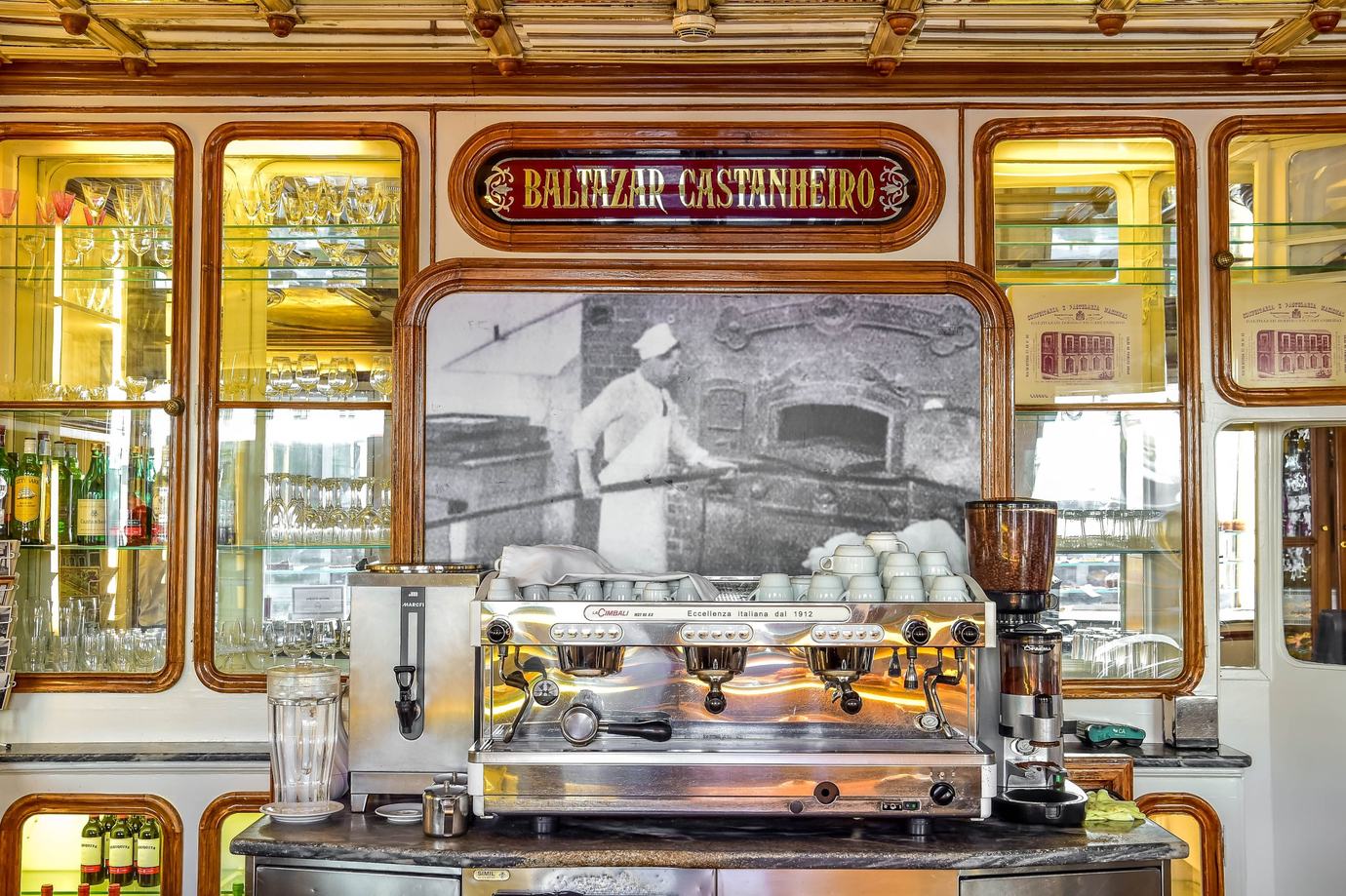 The gilded counter of Confeitaria Nacional in Lisbon, with the founder’s name above an espresso machine and an old photograph of a baker at the oven