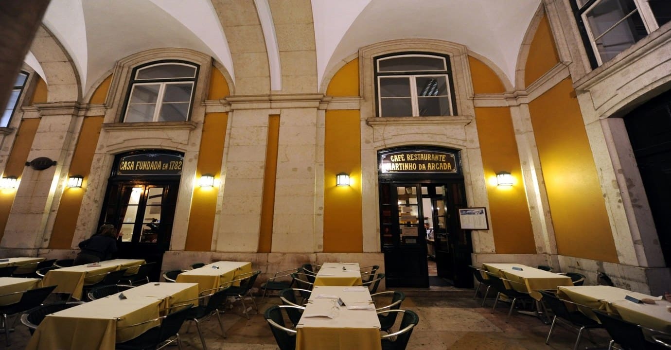 The yellow arcade of Praça do Comércio at night, with the lit entrances of Café Martinho da Arcada and its outdoor tables