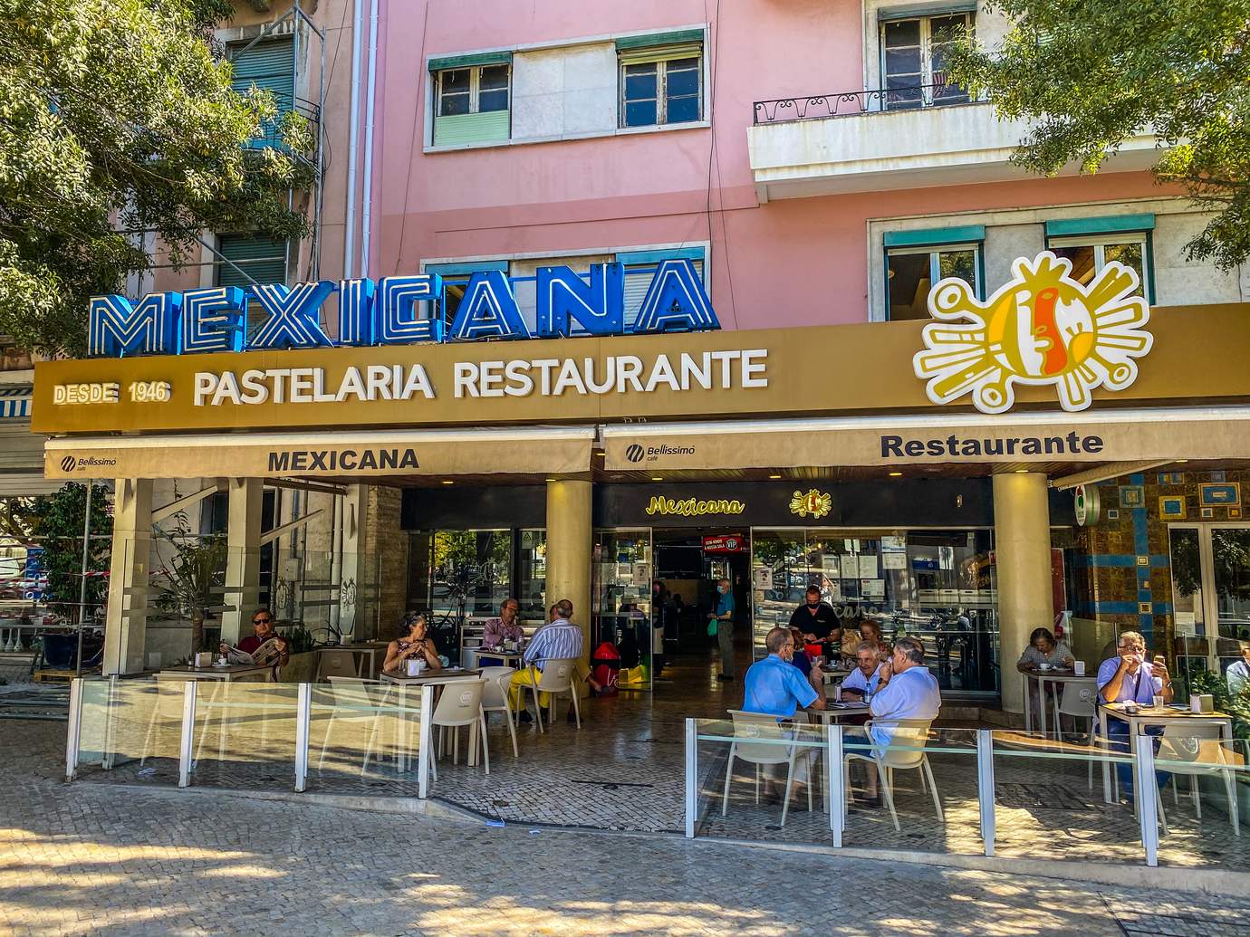The golden sign and sunny terrace of Pastelaria Mexicana on Praça de Londres in Lisbon, with a pink building above