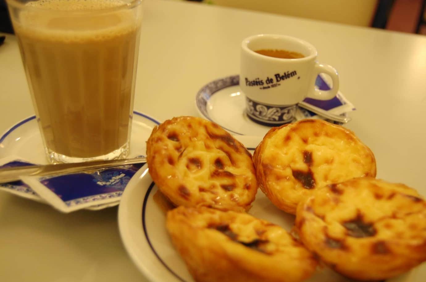Four golden custard tarts and a glass of galão beside an espresso cup printed with Pastéis de Belém