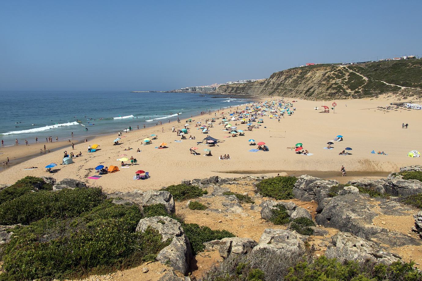 Sunbathers and umbrellas on the wide sandy beach at Foz do Lizandro near Ericeira, with low cliffs and green scrub in the foreground