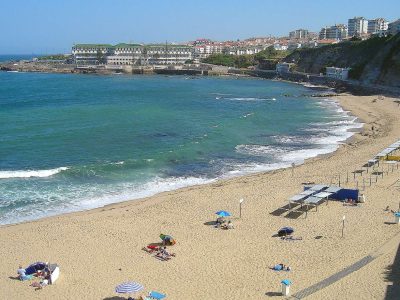 Ericeira Hero Praia Baleia Golden sand and turquoise water at Praia da Baleia in Ericeira, with the town and a large hotel building on the cliff above the bay