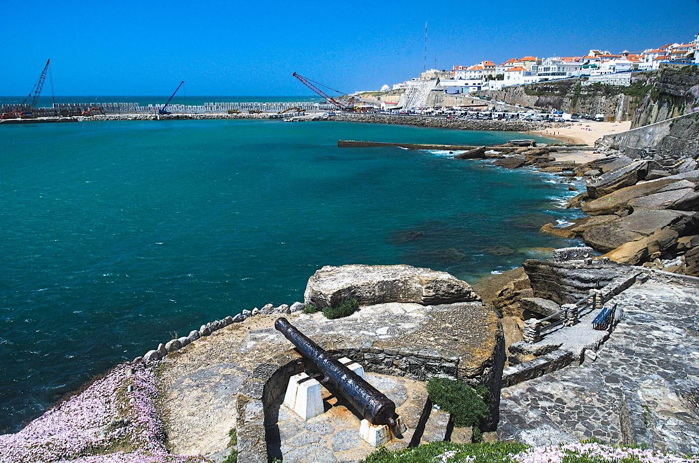 The white houses of Ericeira on a cliff above a turquoise bay, seen from a rocky headland with an old cannon in the foreground