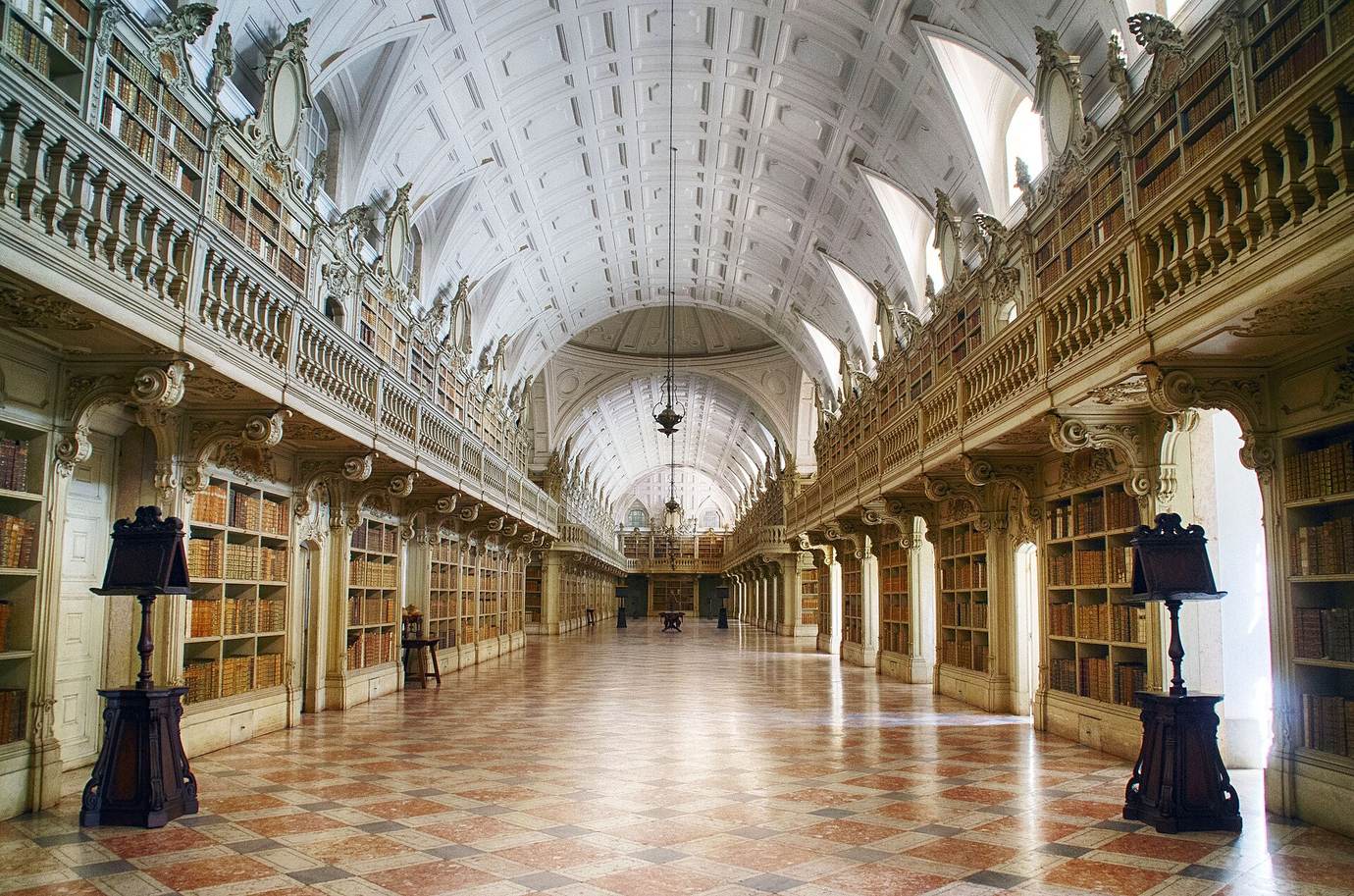 The long baroque library of Mafra Palace with a vaulted white ceiling, carved wooden shelves and a chequered marble floor