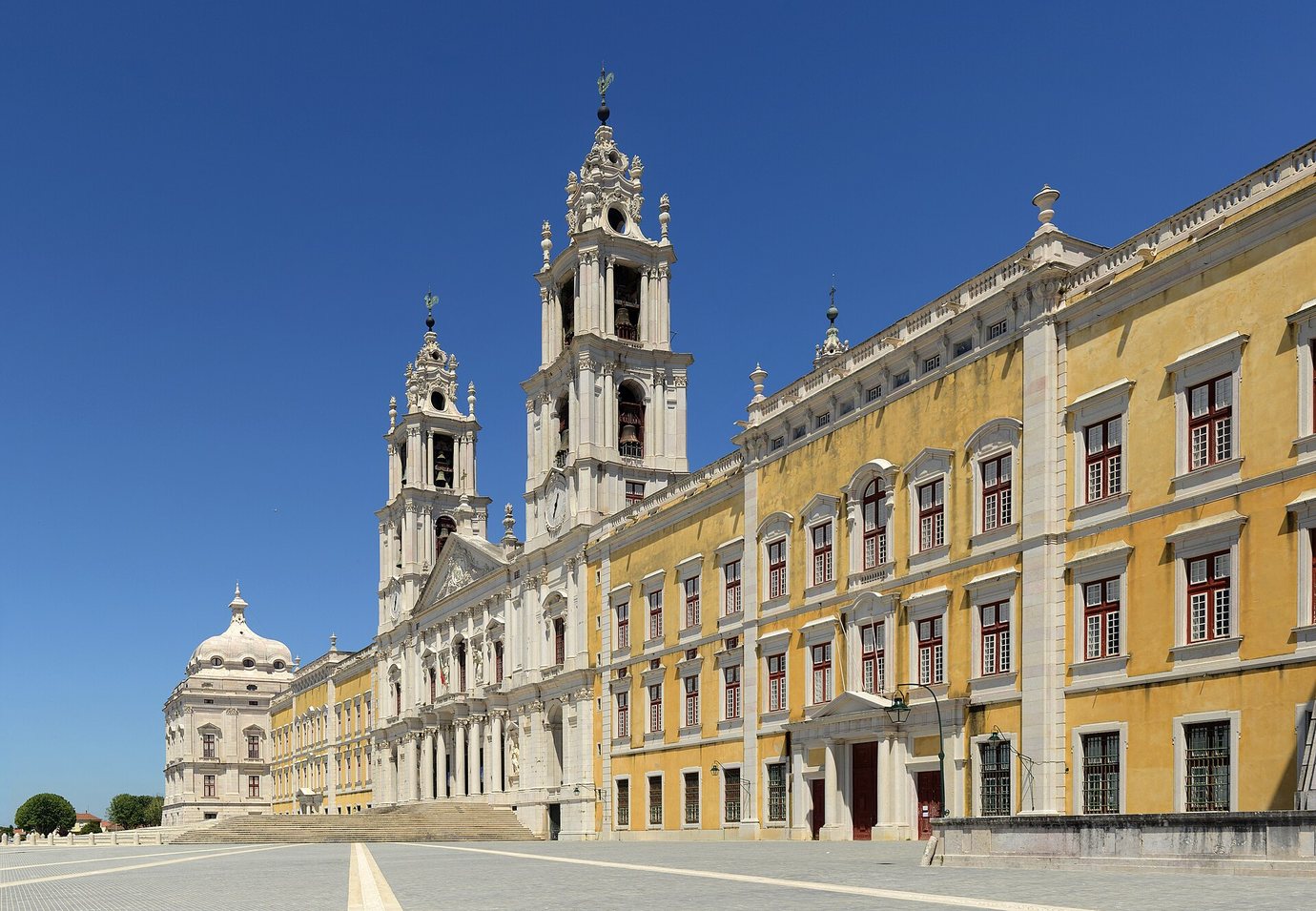 The long yellow and white façade and twin bell towers of the National Palace of Mafra under a deep blue sky