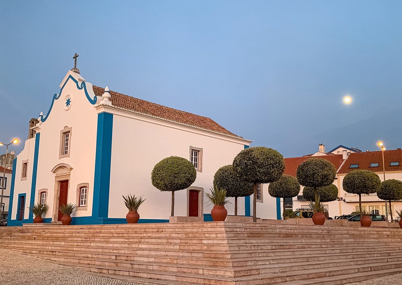 The white and blue baroque church of Santa Marta in Ericeira at dusk, with clipped trees and a broad stepped terrace