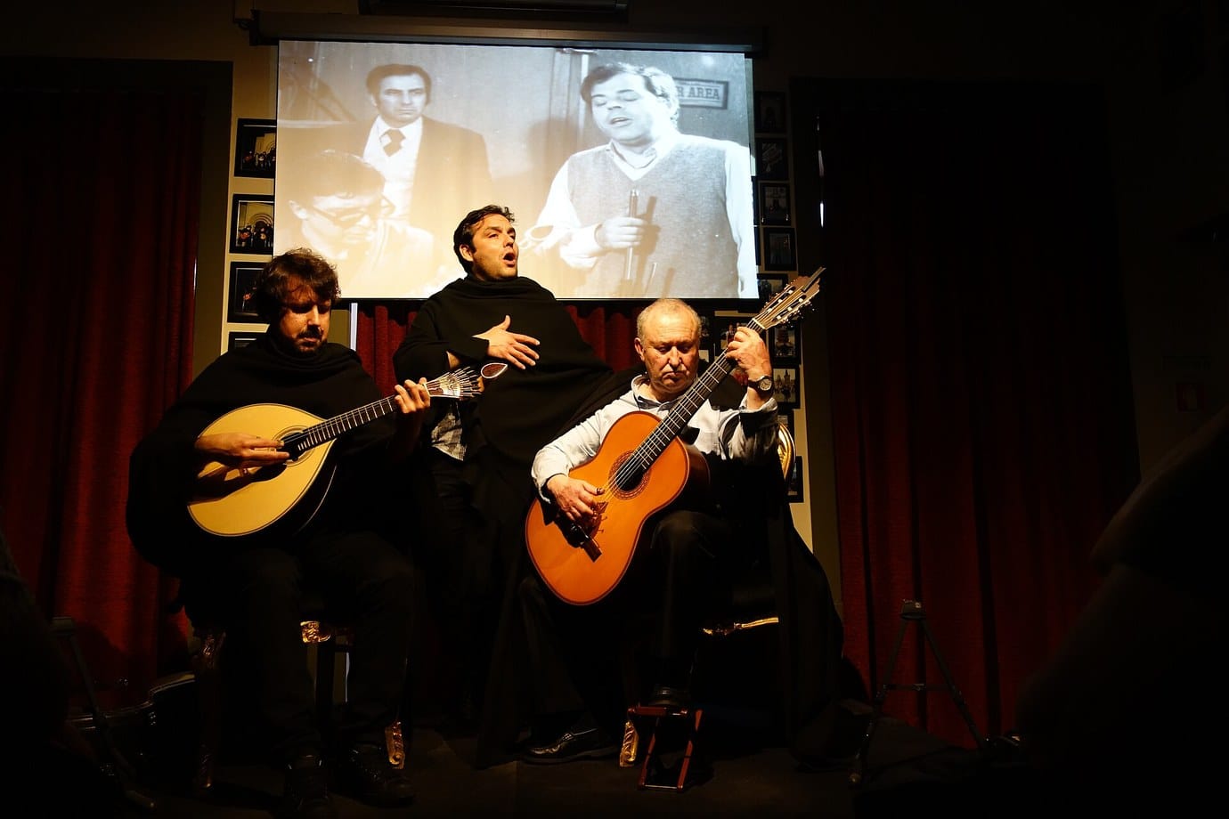 Three men in black capes performing Coimbra fado with a Portuguese guitar and a classical guitar in front of a red curtain