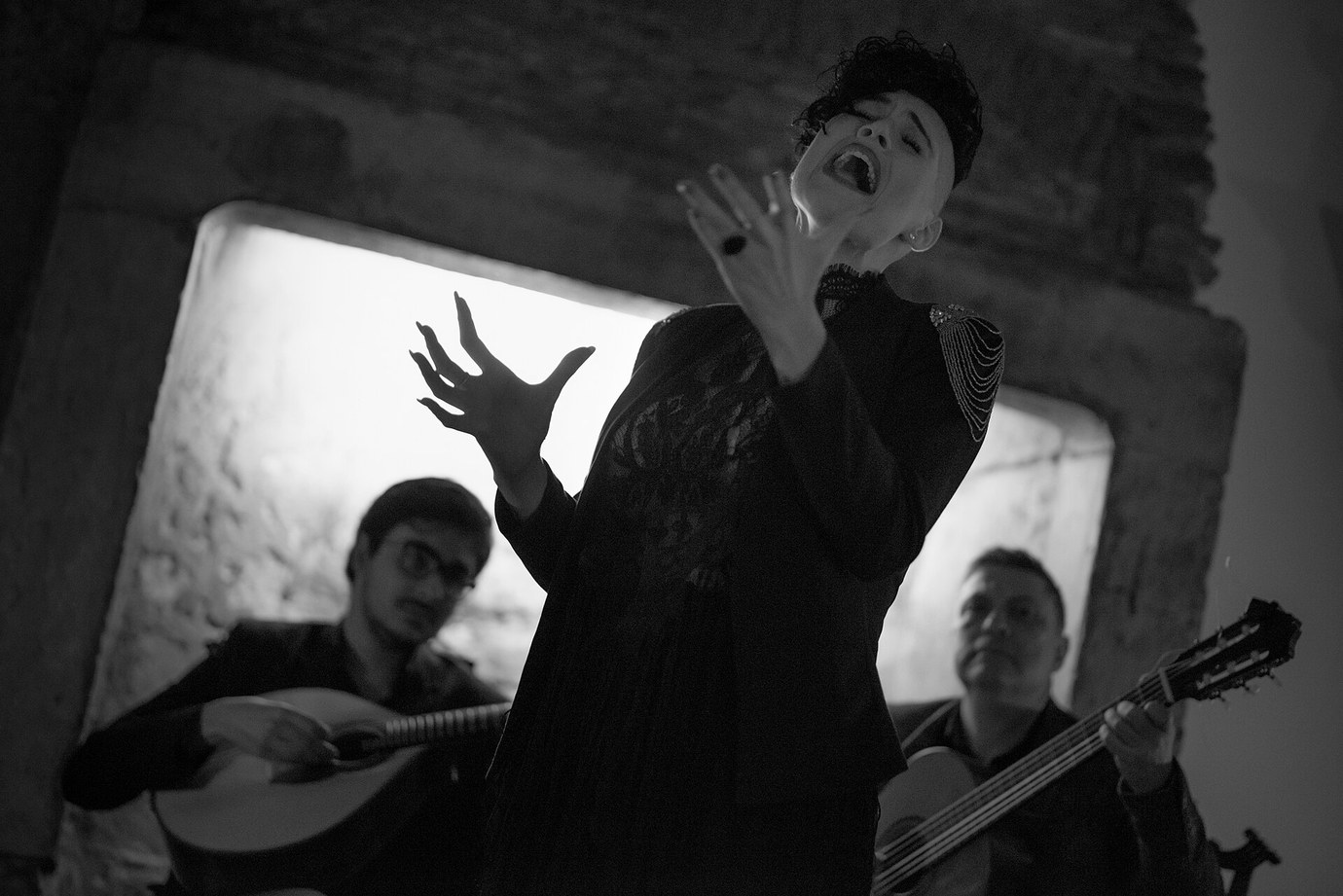 A fadista with her head thrown back and hands raised, framed by a stone window, accompanied by a guitarra portuguesa and a classical guitar