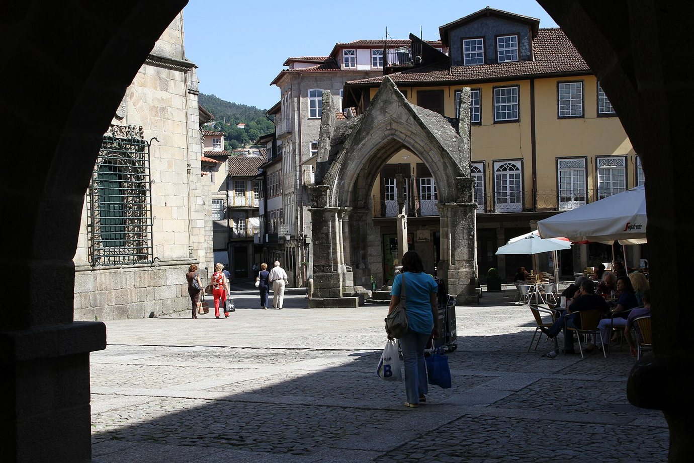 The Gothic Padrão do Salado seen through a granite arch on Largo da Oliveira in Guimarães, with café umbrellas and coloured houses beyond